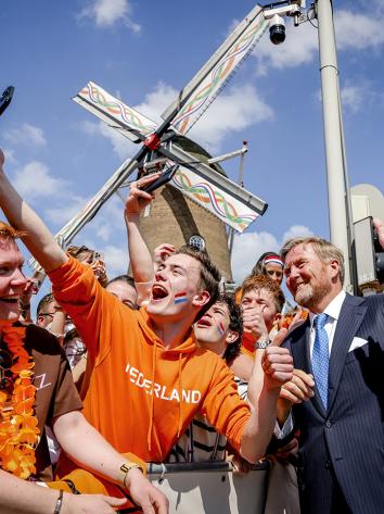 Koning Willem-Alexander maakt een selfie met mensen gekleed in oranje die bij een molen staan achter een hek