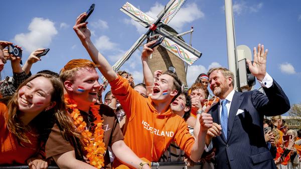 Koning Willem-Alexander maakt een selfie met mensen gekleed in oranje die bij een molen staan achter een hek