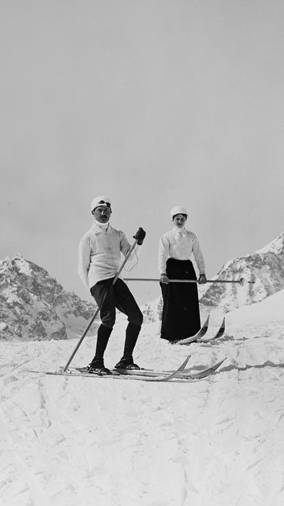 een zwart-witfoto van twee skiërs die eind 19e eeuw poseren voor de camera in de besneeuwde bergtoppen van de Alpen.