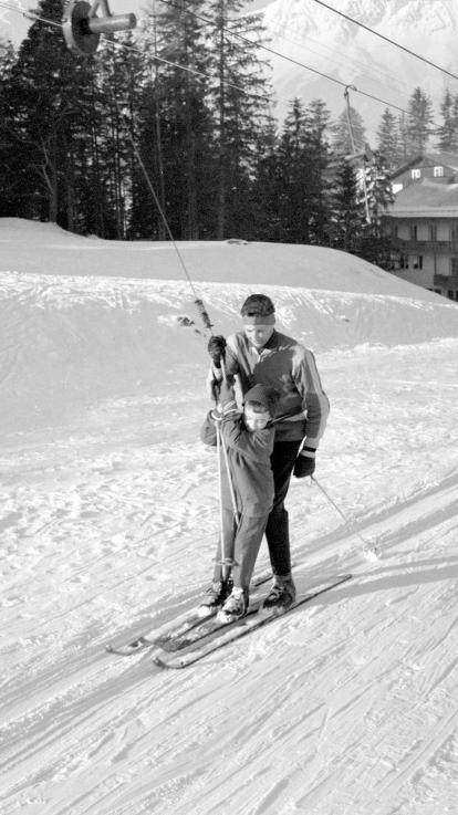 Een zwart-witfoto van ouder en kind die samen op ski's worden voortgetrokken door een sleeplift in de sneeuw