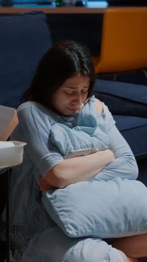 Vrouw zit huilend op de grond voor de bank met armen om een kussen heen geslagen. Ze kijkt omlaag. Naast haar staat een tafeltje voor afval van een maaltijd.