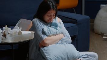 Vrouw zit huilend op de grond voor de bank met armen om een kussen heen geslagen. Ze kijkt omlaag. Naast haar staat een tafeltje voor afval van een maaltijd.