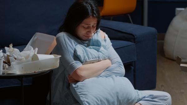 Vrouw zit huilend op de grond voor de bank met armen om een kussen heen geslagen. Ze kijkt omlaag. Naast haar staat een tafeltje voor afval van een maaltijd.