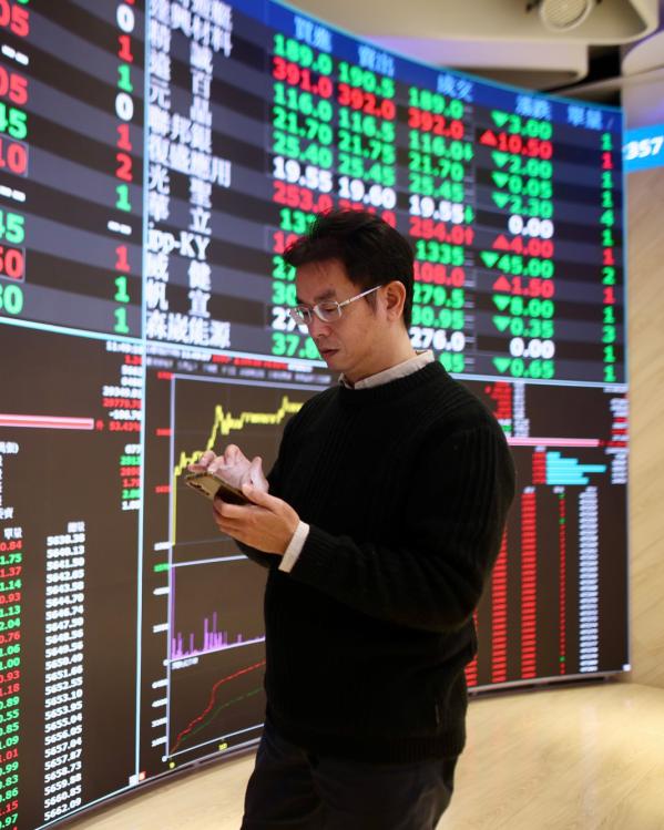 Een man kijkt op zijn mobiel met achter hem een groot scherm met beurskoersen in Taiwan