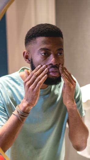 Een man kijkt serieus naar zichzelf in de spiegel terwijl hij met zijn handen zijn gezicht aanraakt.