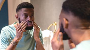 Een man kijkt serieus naar zichzelf in de spiegel terwijl hij met zijn handen zijn gezicht aanraakt.