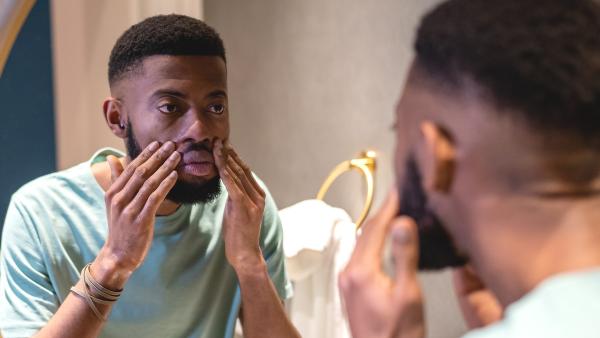 Een man kijkt serieus naar zichzelf in de spiegel terwijl hij met zijn handen zijn gezicht aanraakt.