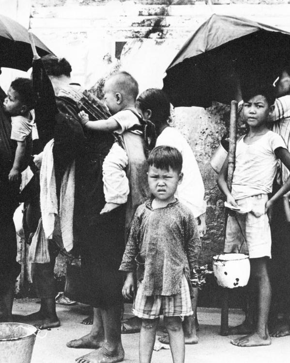 Een zwart-witfoto van mensen met kinderen die in de rij staan voor een maaltijd met hun pan of emmer.