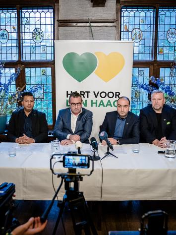 Mannen voor een poster van Hart voor Den Haag zitten aan een tafel met wit tafellaken. Ze worden gefilmd. 