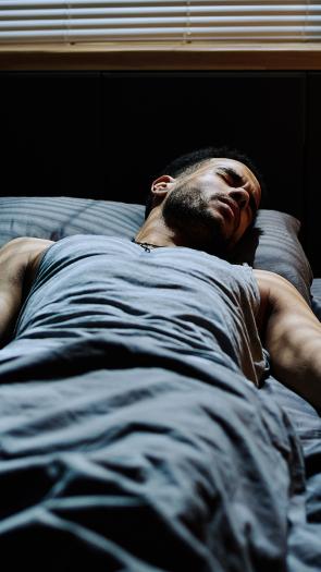 Man ligt in bed en kijkt fronsend in slaap. Er valt licht vanuit raam op zijn lichaam 