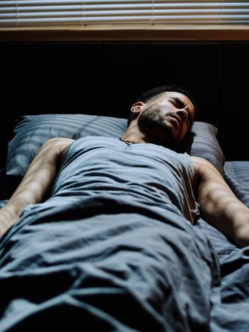 Man ligt in bed en kijkt fronsend in slaap. Er valt licht vanuit raam op zijn lichaam 