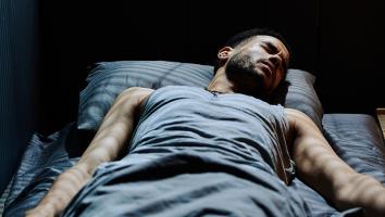 Man ligt in bed en kijkt fronsend in slaap. Er valt licht vanuit raam op zijn lichaam 