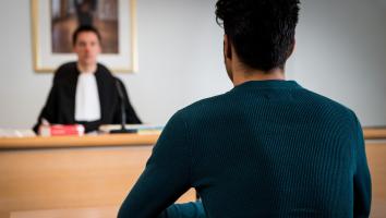 Rug van een jongen in de rechtszaal. Onscherp zit er een rechter tegenover hem. 