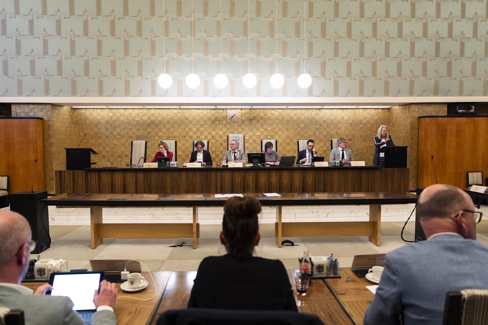 op een rijtje aan een houten tafel zit de burgemeester van Amersfoort naast de wethouders. Voor zie je de rug van drie raadsleden van de gemeenteraad. 