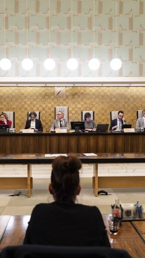 op een rijtje aan een houten tafel zit de burgemeester van Amersfoort naast de wethouders. Voor zie je de rug van drie raadsleden van de gemeenteraad. 