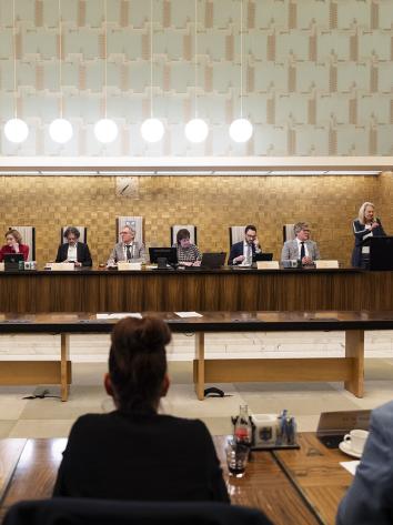 op een rijtje aan een houten tafel zit de burgemeester van Amersfoort naast de wethouders. Voor zie je de rug van drie raadsleden van de gemeenteraad. 