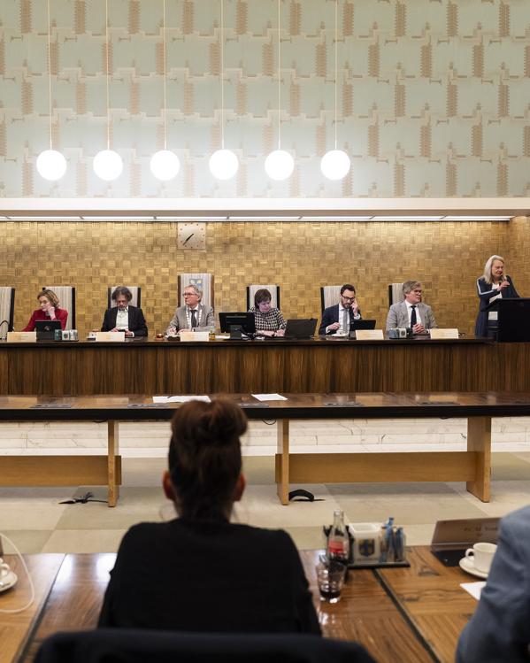 op een rijtje aan een houten tafel zit de burgemeester van Amersfoort naast de wethouders. Voor zie je de rug van drie raadsleden van de gemeenteraad. 