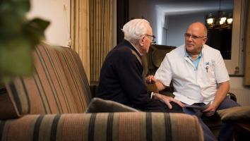 Een oude man praat met een kale man in doktersuniform. Ze zitten in een woonkamer op een oude bank. 