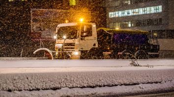 Een sneeuwschuiver rijdt door een straat vol sneeuw met zwaailichten aan. Er valt ook sneeuw uit de lucht