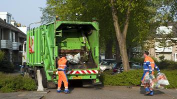 Een vuilnisman gooit zakken afval in een vuilniswagen. Rechts komt nog een vuilnisman aangelopen met twee zakken vast.