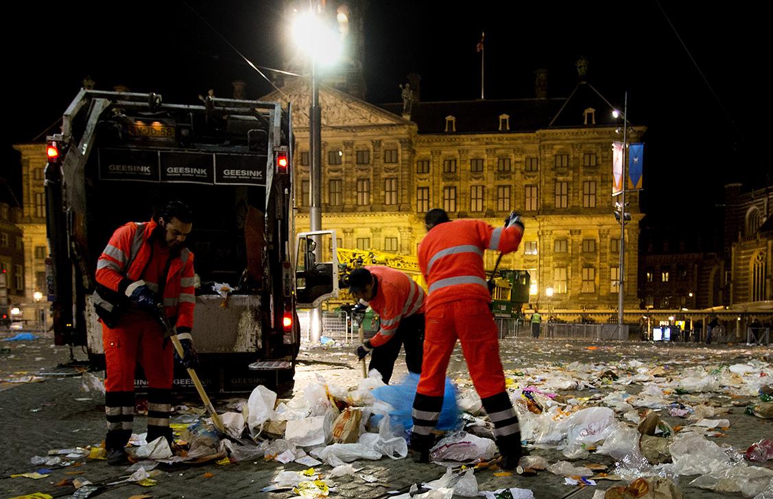 Drie vuilnismannen zijn 's nachts een plein aan het opruimen waar veel plastic en afval op straat ligt.