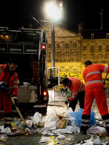 Drie vuilnismannen zijn 's nachts een plein aan het opruimen waar veel plastic en afval op straat ligt.
