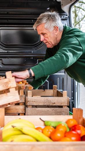 een man met grijze haren aan het werk, hij pakt kratten met fruit en groente uit een busje