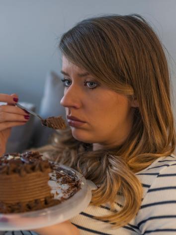 Een vrouw zit op de bank en kijkt verdrietig voor zich uit terwijl ze een stukje chocoladetaart eet. Ze houdt de rest van de taart vast op een plateautje in haar andere hand.