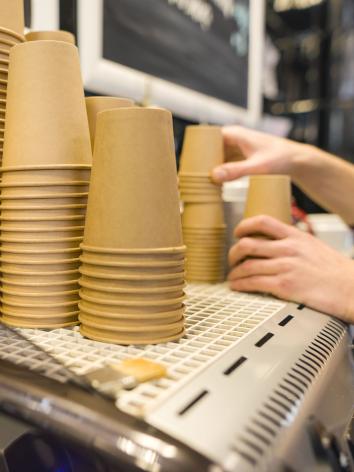 Op een groot koffiezetapparaat staan stapels bruine, kartonnen koffiebekers. Twee handen reiken naar de bekers.