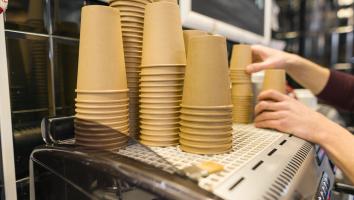 Op een groot koffiezetapparaat staan stapels bruine, kartonnen koffiebekers. Twee handen reiken naar de bekers.