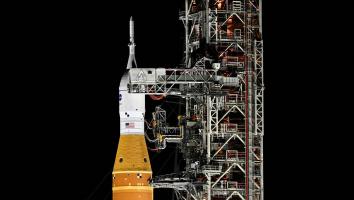 De Space Launch System-raket met het Orion-ruimteschip staat rechtop op het lanceerplatform, naast een hoge metalen lanceertoren, verlicht in de nacht.
