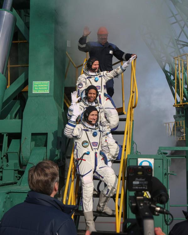De astronauten staan op een rijtje op de trap te zwaaien naar de pers. Op de achtergrond zie je delen van de lanceerinstallatie.