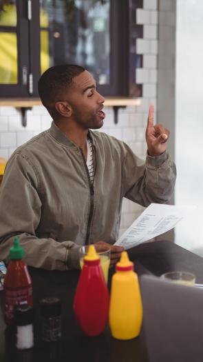 Man en vrouw aan tafel in restaurant. Man bestelt bij serveerster met menu in hand en vinger in de lucht gestoken.