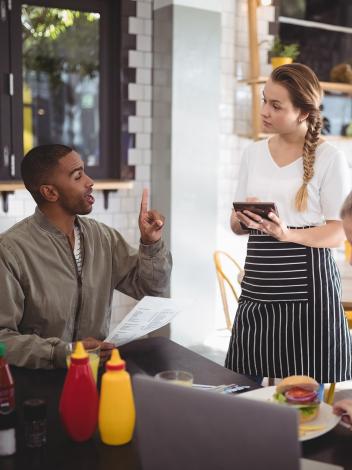 Man en vrouw aan tafel in restaurant. Man bestelt bij serveerster met menu in hand en vinger in de lucht gestoken.