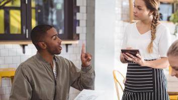 Man en vrouw aan tafel in restaurant. Man bestelt bij serveerster met menu in hand en vinger in de lucht gestoken.