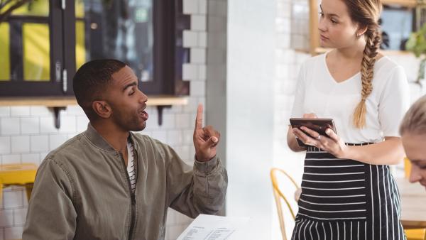 Man en vrouw aan tafel in restaurant. Man bestelt bij serveerster met menu in hand en vinger in de lucht gestoken.