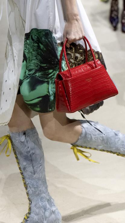 Een model loopt met een kleine rode tas, hoge grijze laarzen met gele veters en een wit/groene jurk.