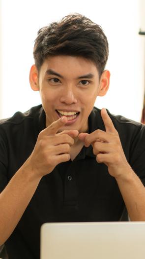 Een jongvolwassen man wijst met beide vingers naar de videocamera terwijl hij iets aan het vertellen is.