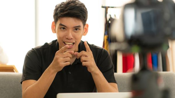 Een jongvolwassen man wijst met beide vingers naar de videocamera terwijl hij iets aan het vertellen is.