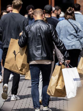 Een man loop in een winkelstraat. Vanaf de achterkant zien we dat hij veel verschillende winkeltassen vast heeft.