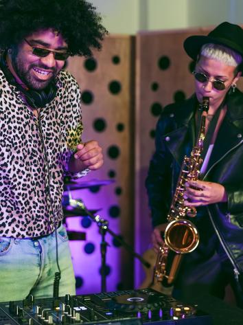 Man met afro en zonnebril en geprinte blouse maakt muziek op dj-set met daarnaast een vrouw die saxofoon speelt