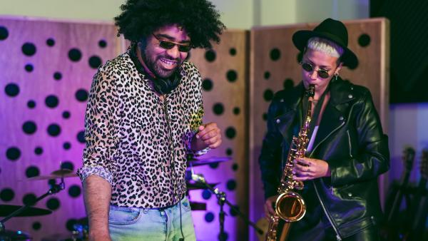 Man met afro en zonnebril en geprinte blouse maakt muziek op dj-set met daarnaast een vrouw die saxofoon speelt