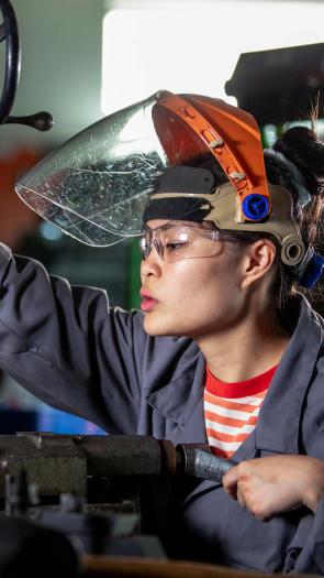 Vrouw met oogbescherming en werkkleding boort een gat in een buis bij een techniekwerkplaats
