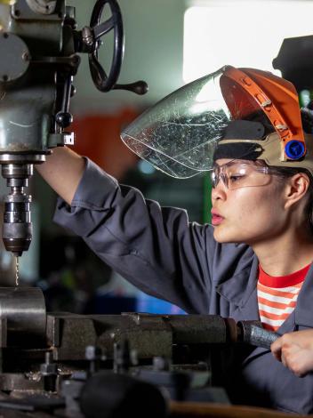 Vrouw met oogbescherming en werkkleding boort een gat in een buis bij een techniekwerkplaats