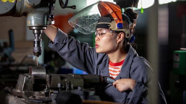 Vrouw met oogbescherming en werkkleding boort een gat in een buis bij een techniekwerkplaats
