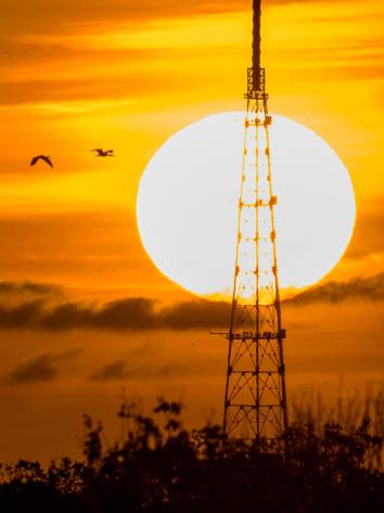 Een opkomende zon met een oranje gloed in de lucht en op de voorgrond silhouetten van een raketinstallatie van NASA