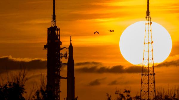 Een opkomende zon met een oranje gloed in de lucht en op de voorgrond silhouetten van een raketinstallatie van NASA