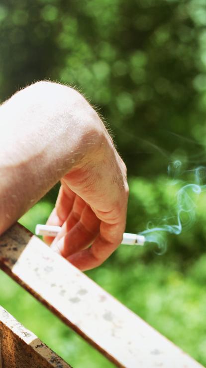 Hand met dampende sigaret hangt over een balkonrand met bomen op de achtergrond