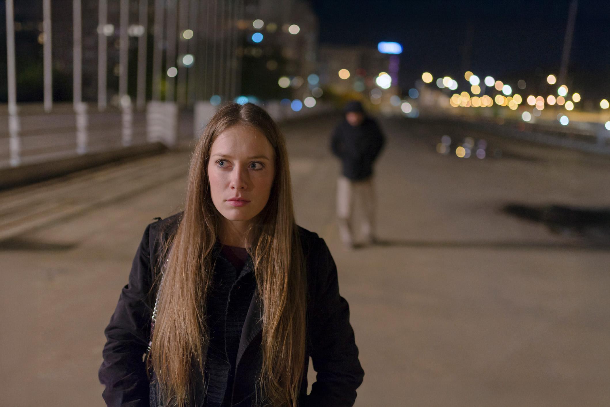 Meisje loopt over een weg in het donker en kijkt bezorgd. Achter haar loopt een man met capuchon onscherp in beeld