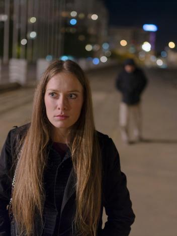 Meisje loopt over een weg in het donker en kijkt bezorgd. Achter haar loopt een man met capuchon onscherp in beeld